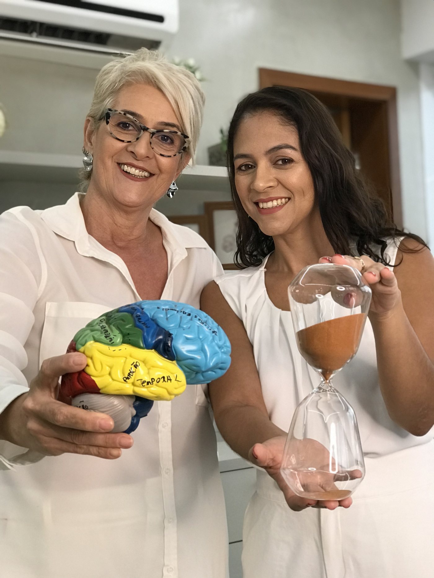 Elaine Castilho segurando um modelo anatômico de cérebro e Cinthia Barbosa segurando uma ampulheta
